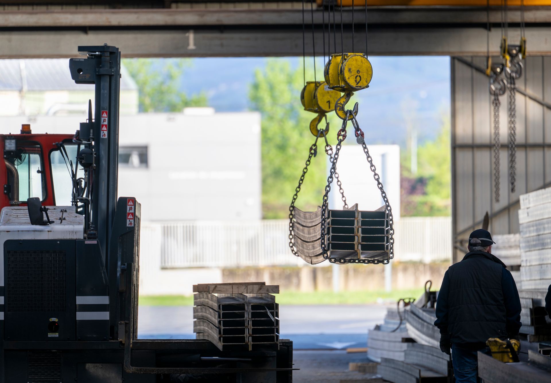 An overhead crane lifting stacked steel beams inside a warehouse while a worker walks nearby. An overhead crane lifting stacked steel beams inside a warehouse while a worker walks nearby.