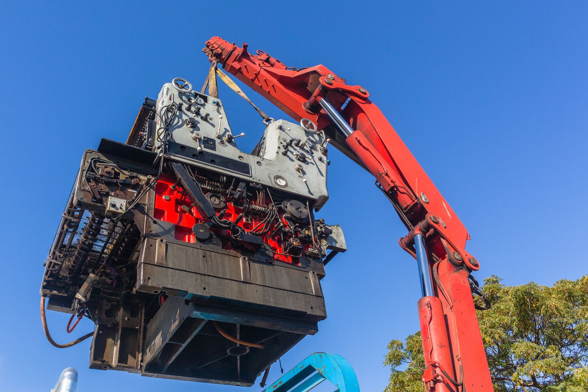A crane is lifting a machine using a spreader bar with a blue sky in the background. A crane is lifting a machine using a spreader bar with a blue sky in the background.