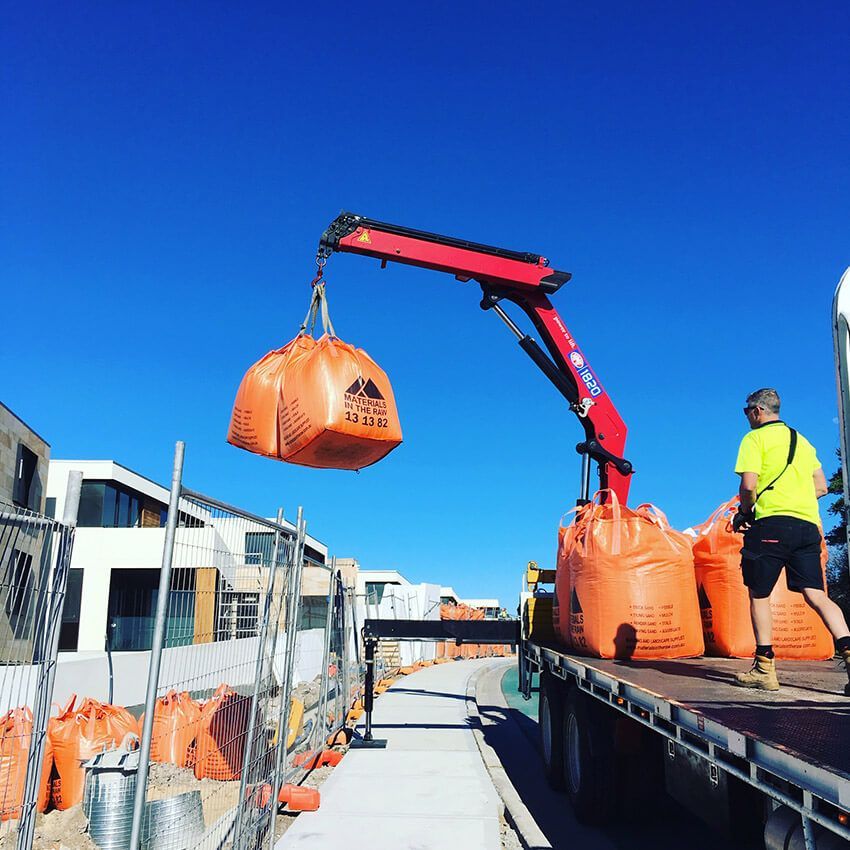 A truck-mounted crane lifts an orange bag at a construction site.