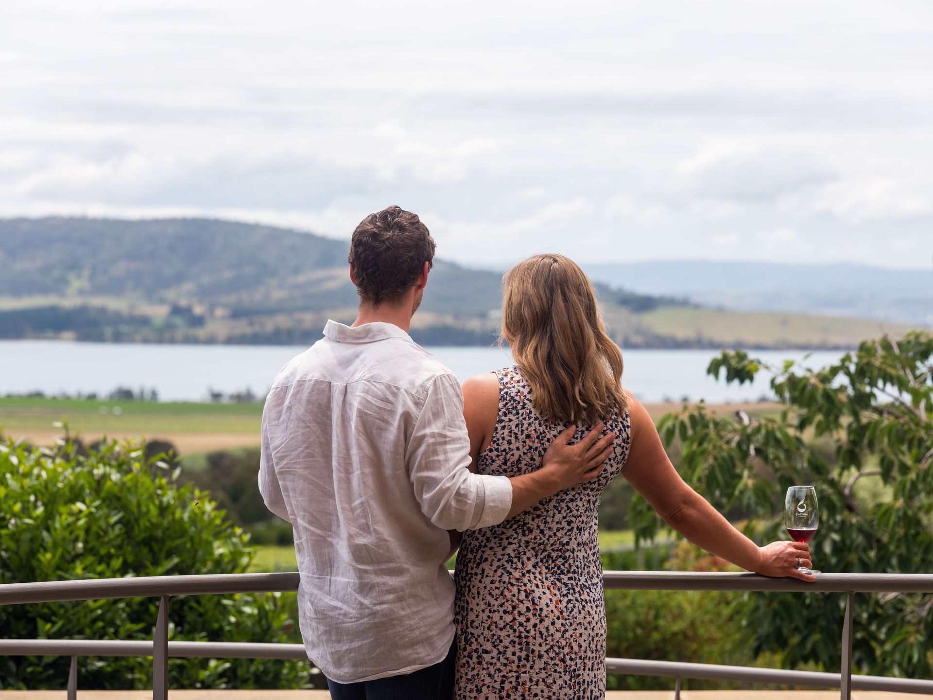 A man and a woman are standing on a balcony looking out over a lake.