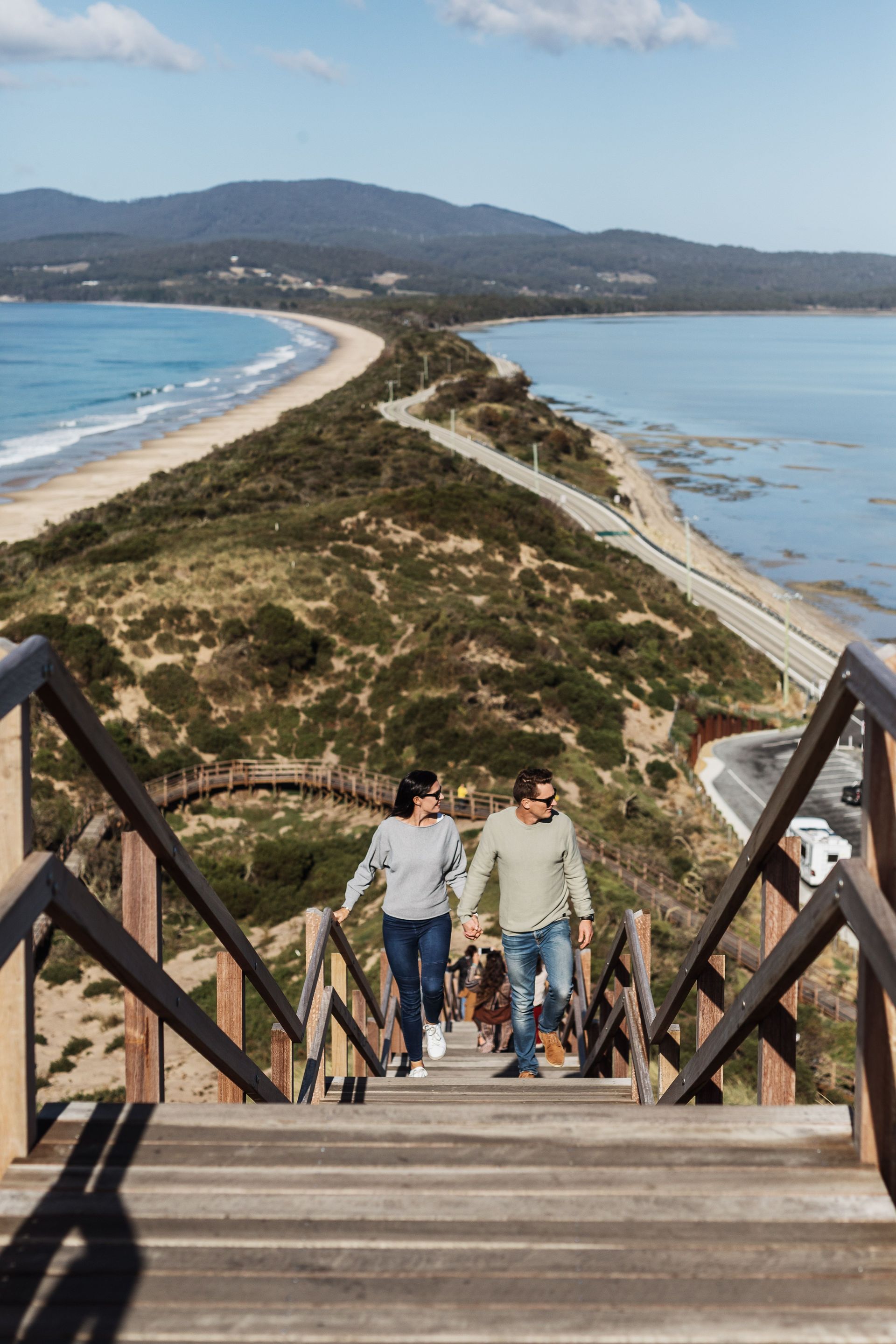 A man and a woman are walking down wooden stairs towards the ocean.