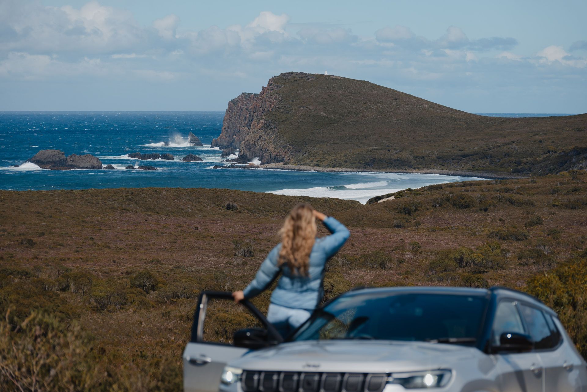 A woman is standing next to a car on a hill overlooking the ocean.
