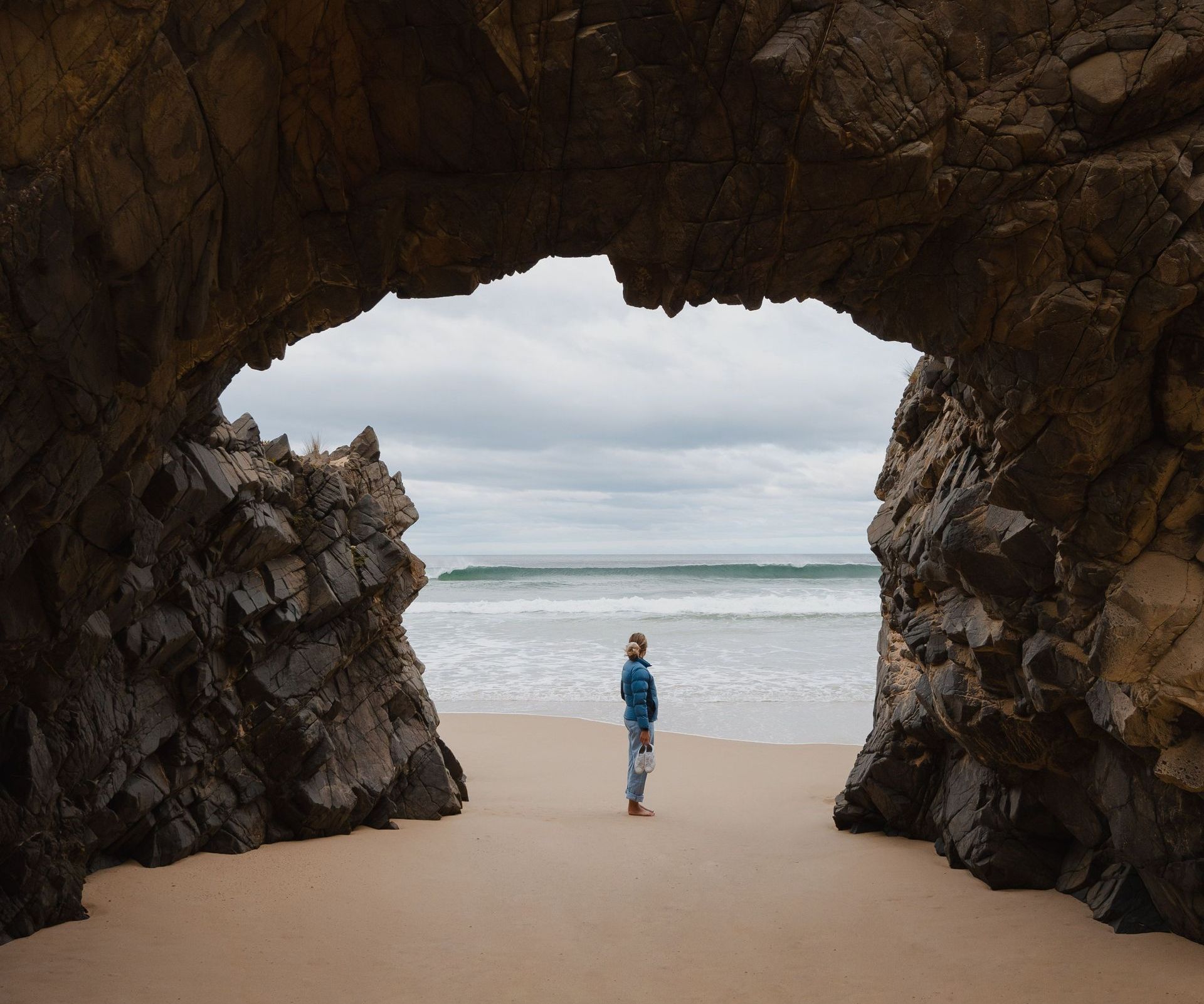 A man is standing in a cave on the beach looking at the ocean.
