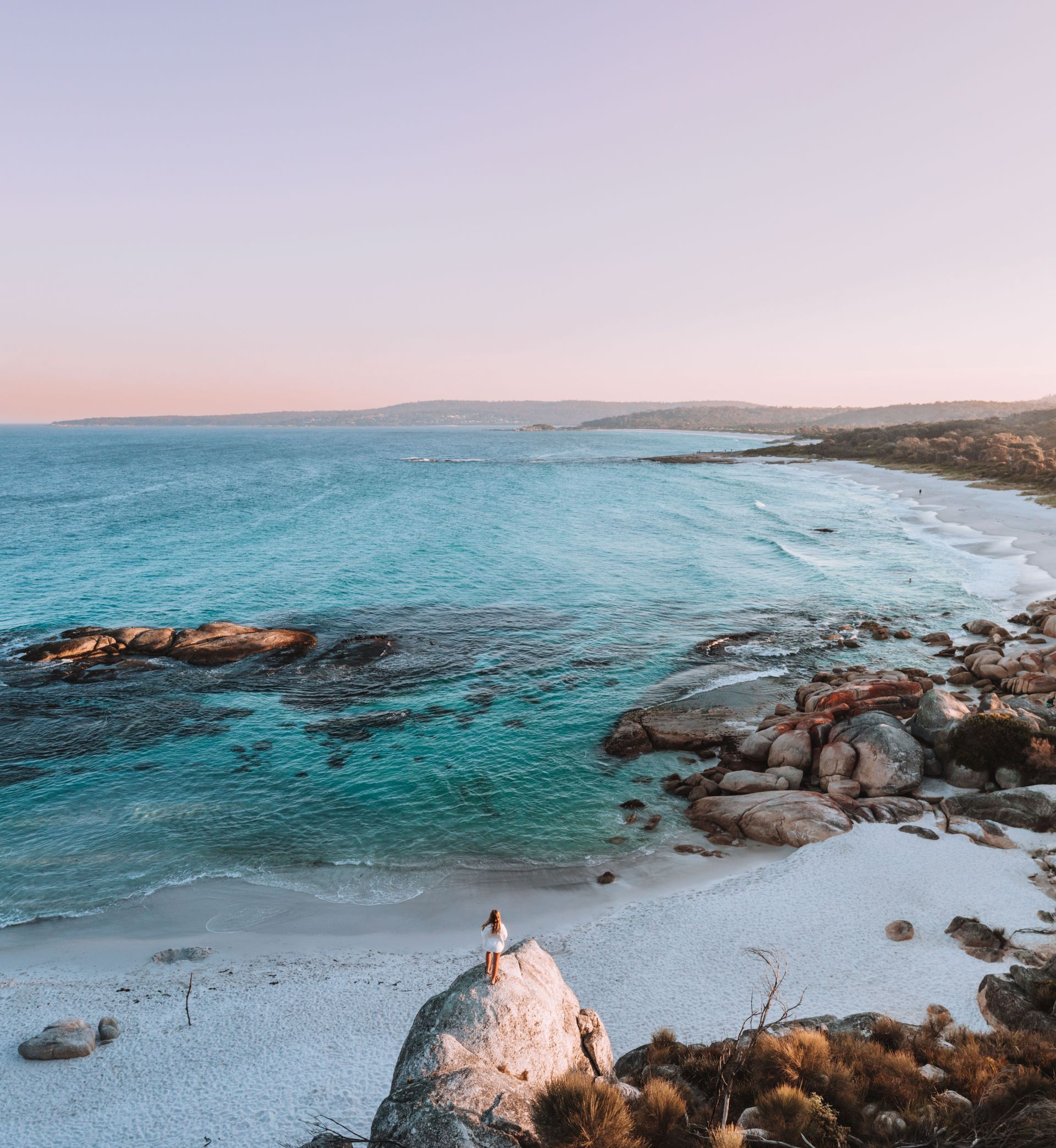 A woman is standing on a rock on a beach overlooking the ocean.