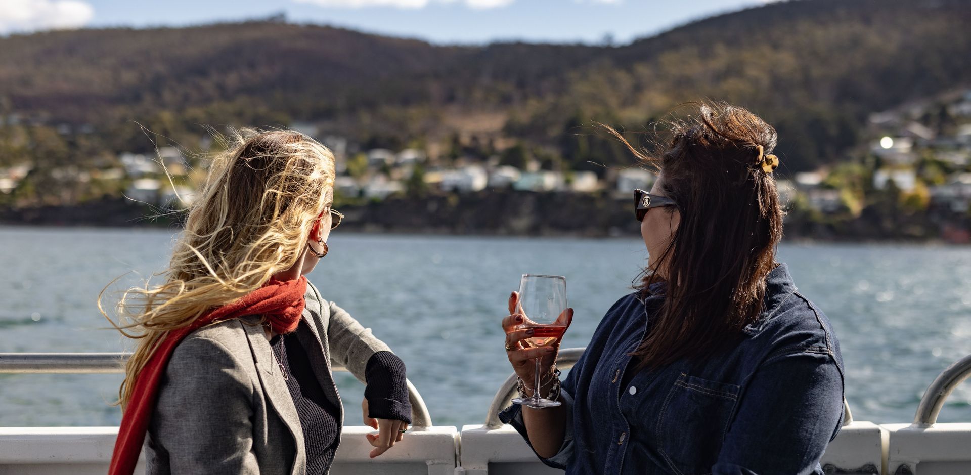 Two women are standing on a boat looking at the water . one of the women is holding a glass of wine.