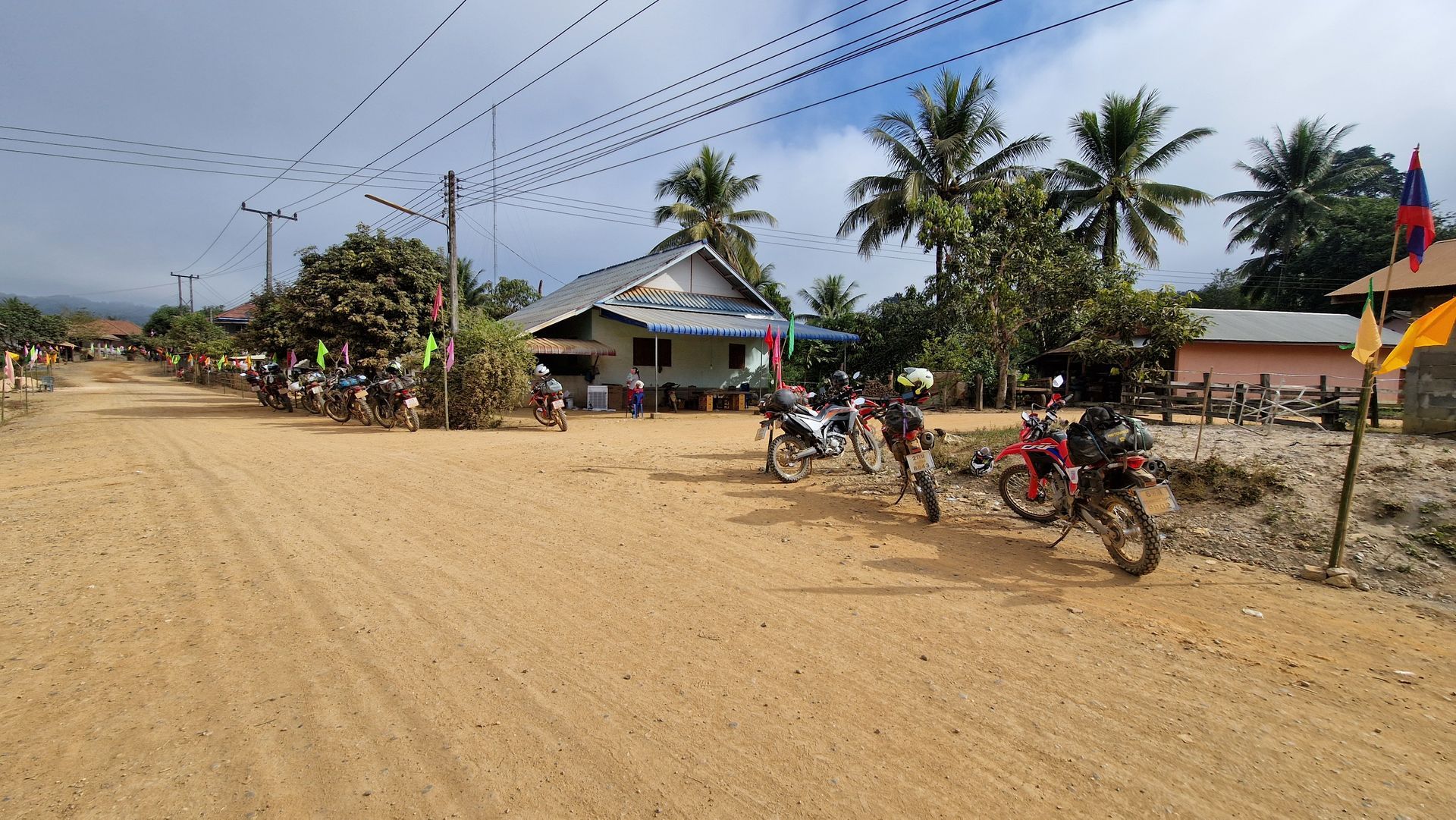A dirt road in a village with several parked motorbikes, palm trees, and houses under a partly cloudy blue sky.