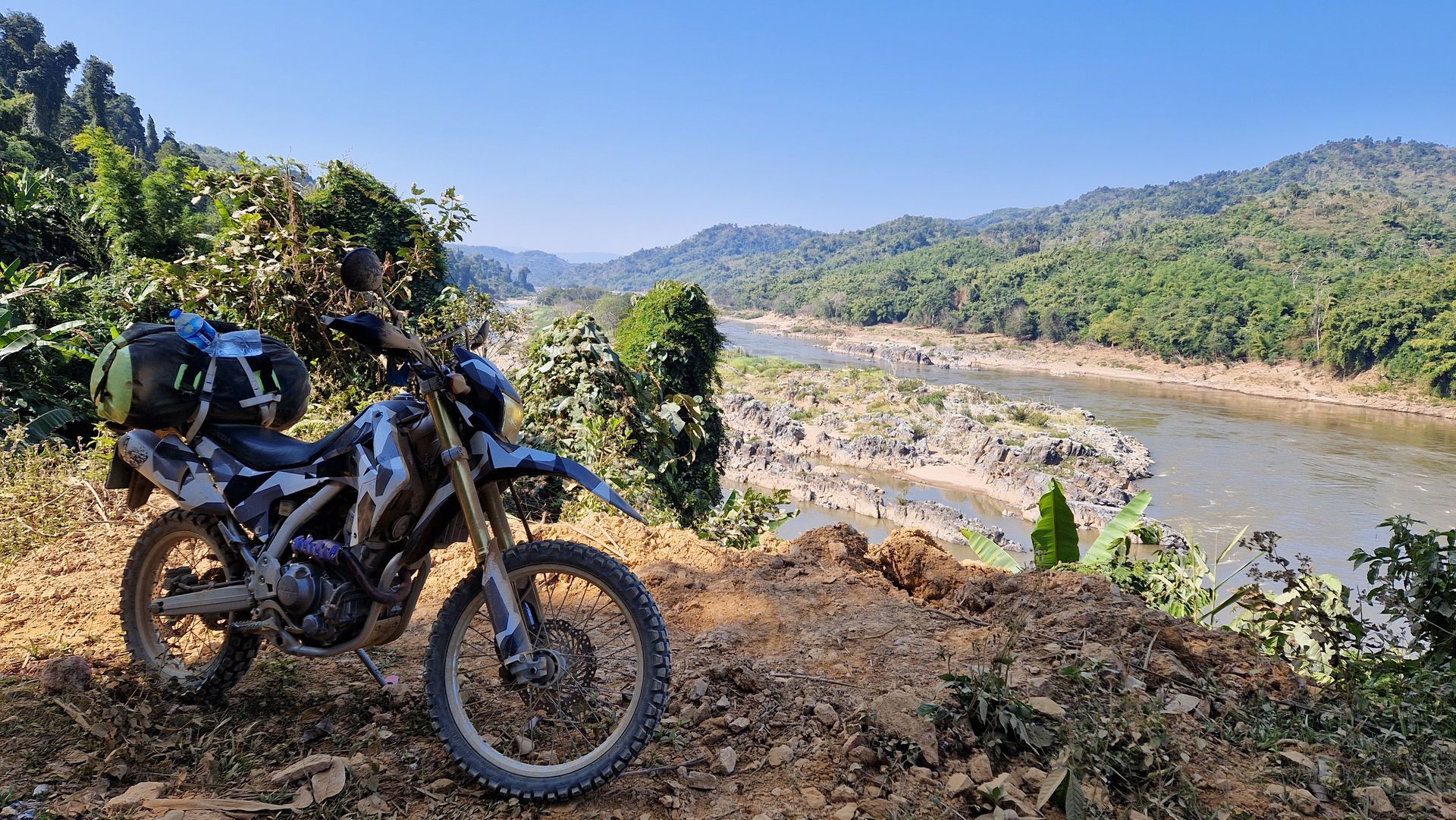 A dual-sport motorcycle with gear strapped to the back, parked on a dirt trail overlooking a river in a forested valley.