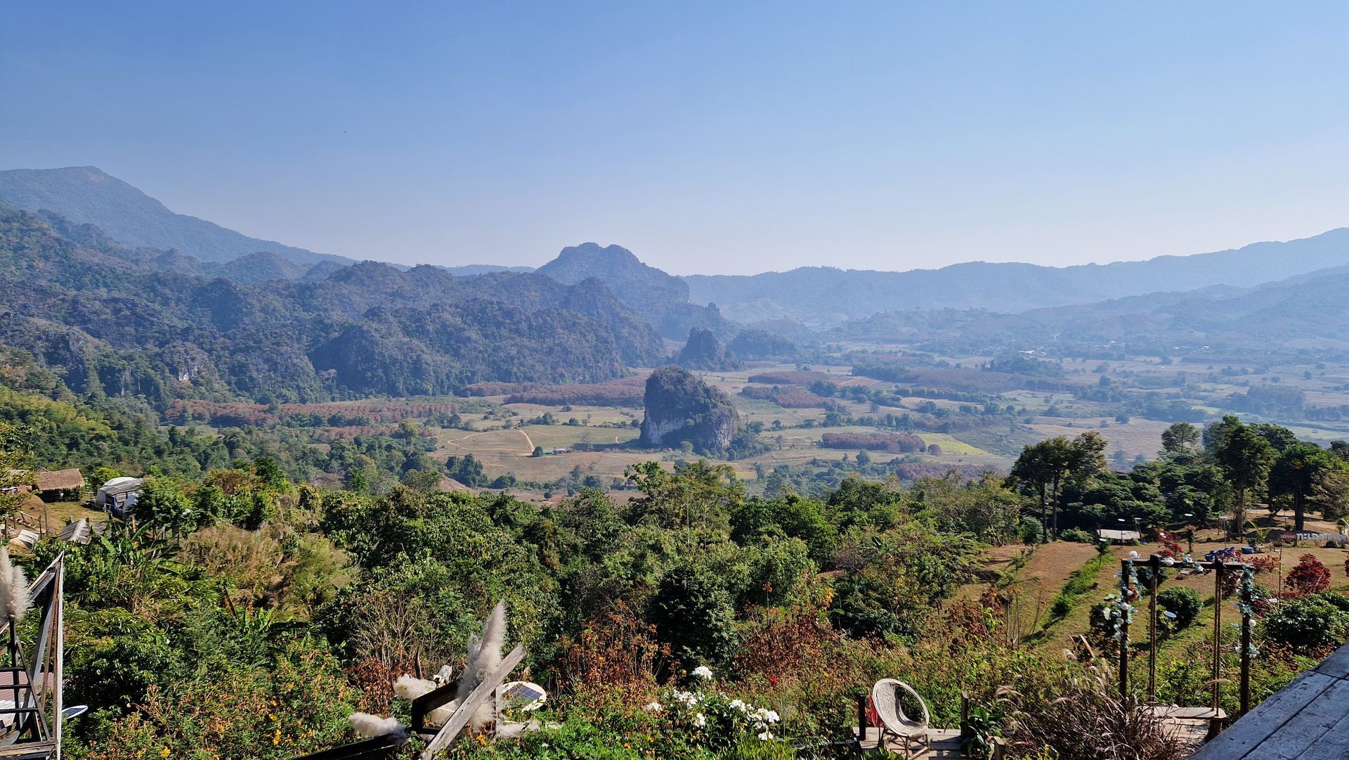 A wide, sunny mountain landscape featuring a distinctive lone rock formation rising from a valley of trees and fields.