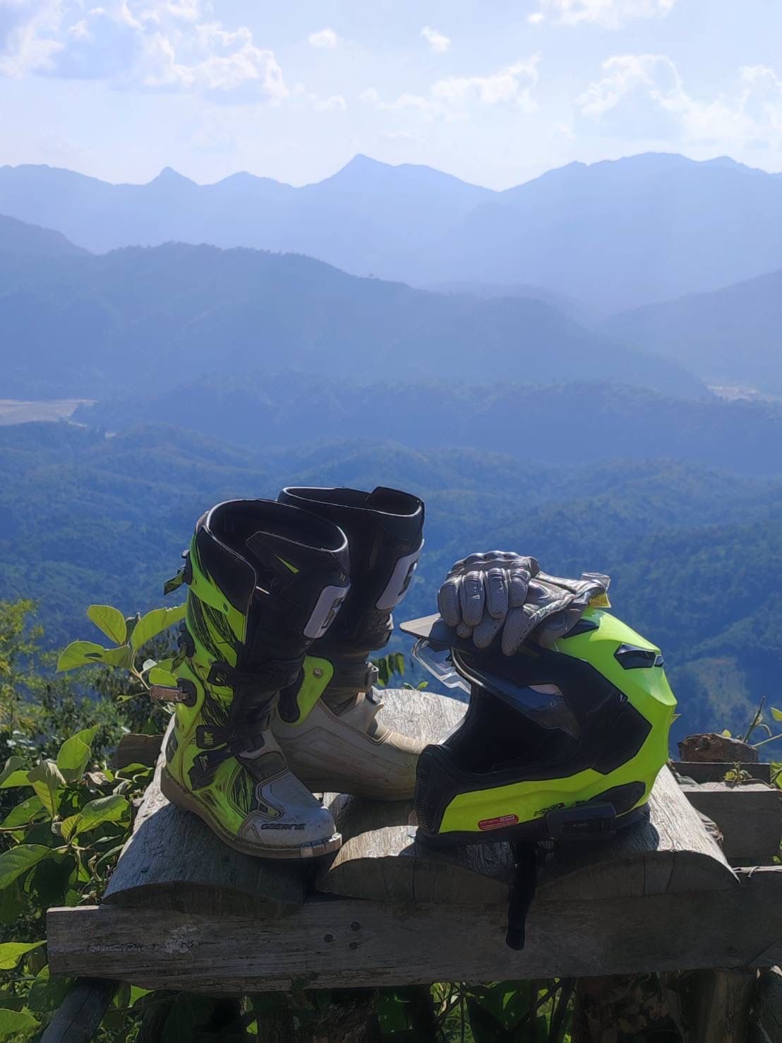 Motocross boots, gloves, and a yellow helmet resting on a wooden platform overlooking a vast mountain range.
