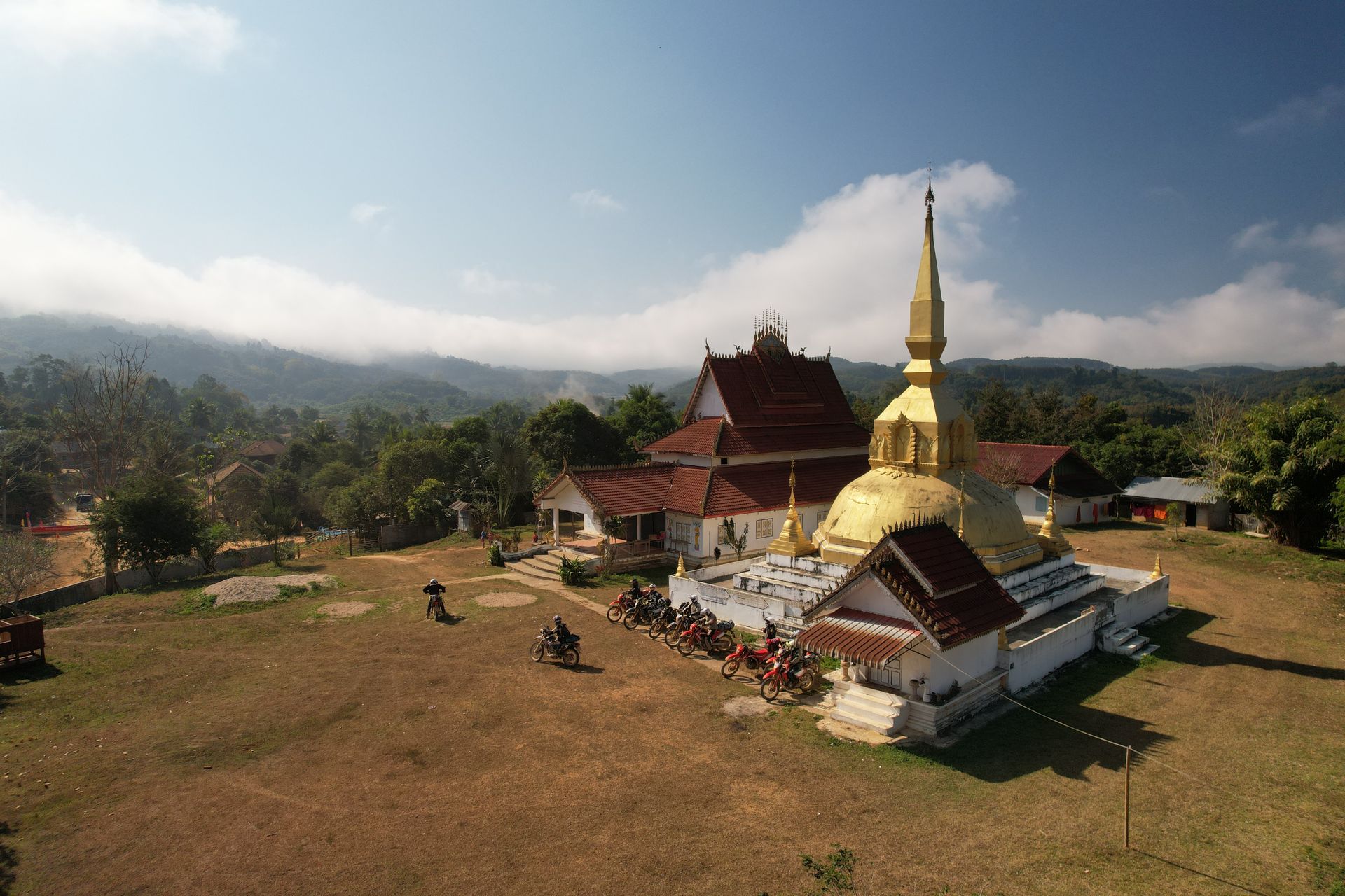 A golden pagoda and temple with tiered, dark-red roofs stand on a dry, grassy field against a backdrop of cloudy mountains. Off road in Laos.