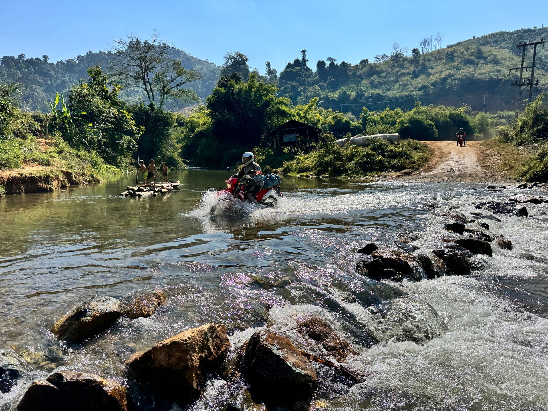 A rider on dirt bike crosses a rocky, shallow river in a lush, mountainous landscape under a bright, clear blue sky.