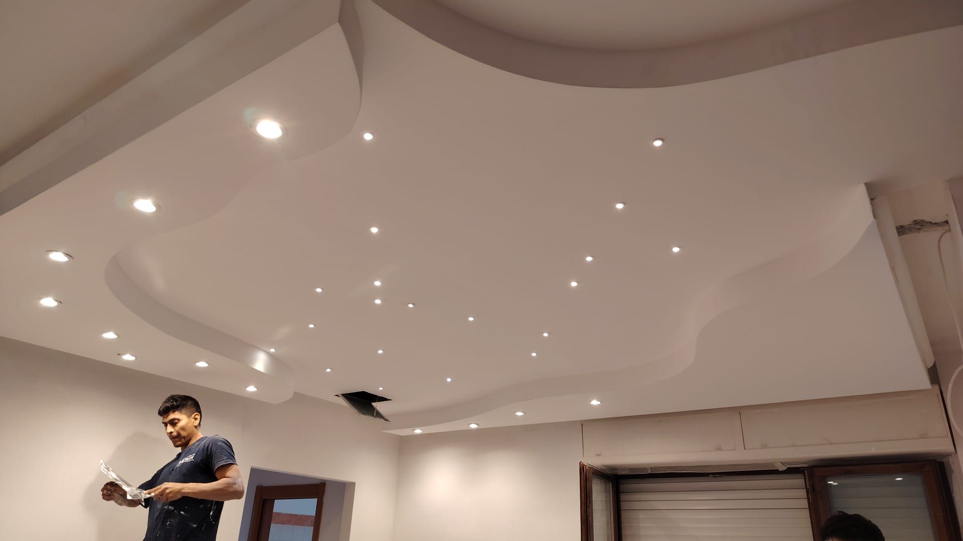 Uomo in piedi in una stanza moderna con soffitto bianco curvo e faretti a incasso, che tiene in mano una macchina fotografica o un dispositivo.
