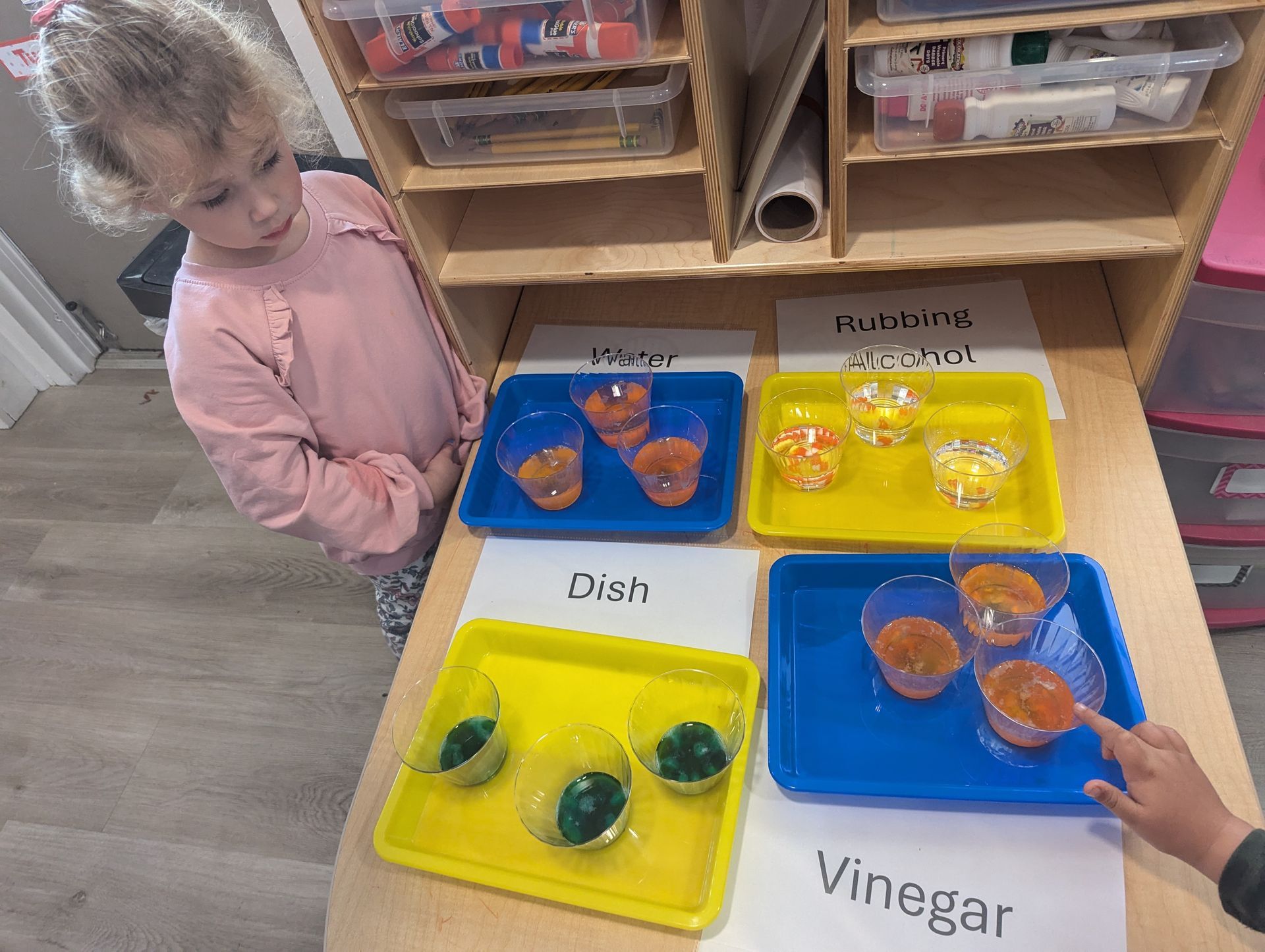 A little girl is standing at a table with trays of colored liquids.