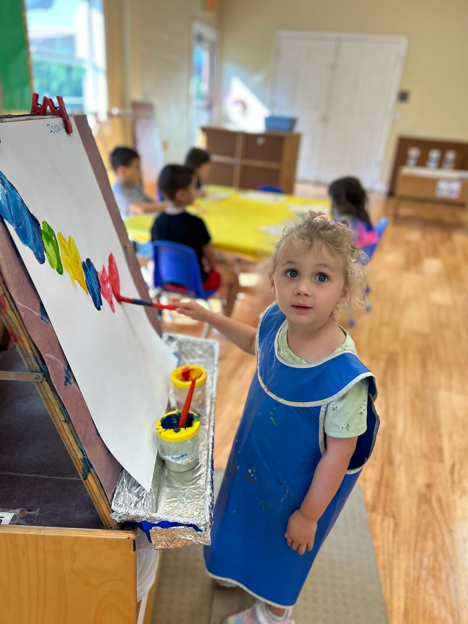 A little girl in a blue apron is painting on an easel.