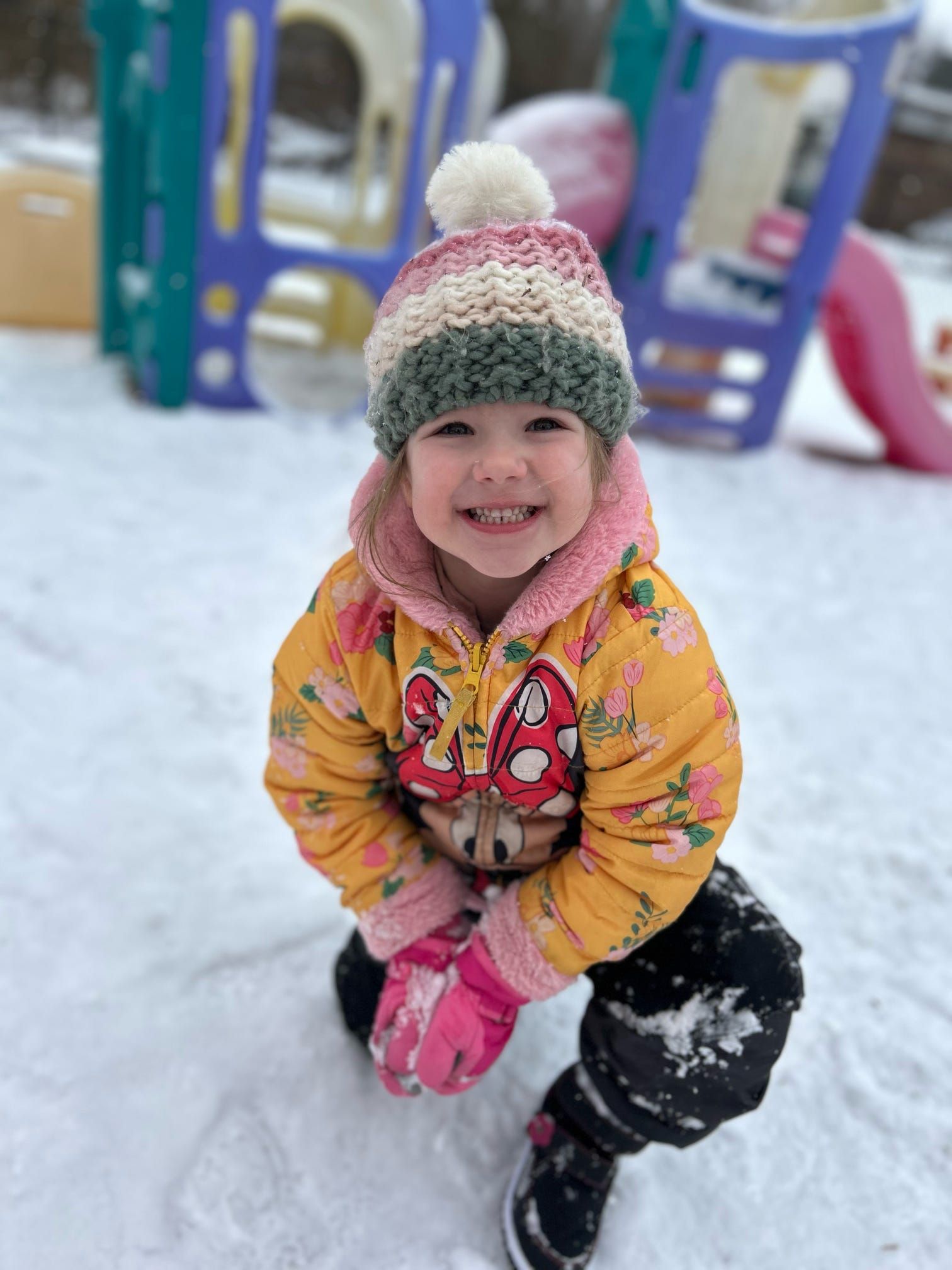 A little girl wearing a hat and gloves is kneeling in the snow.
