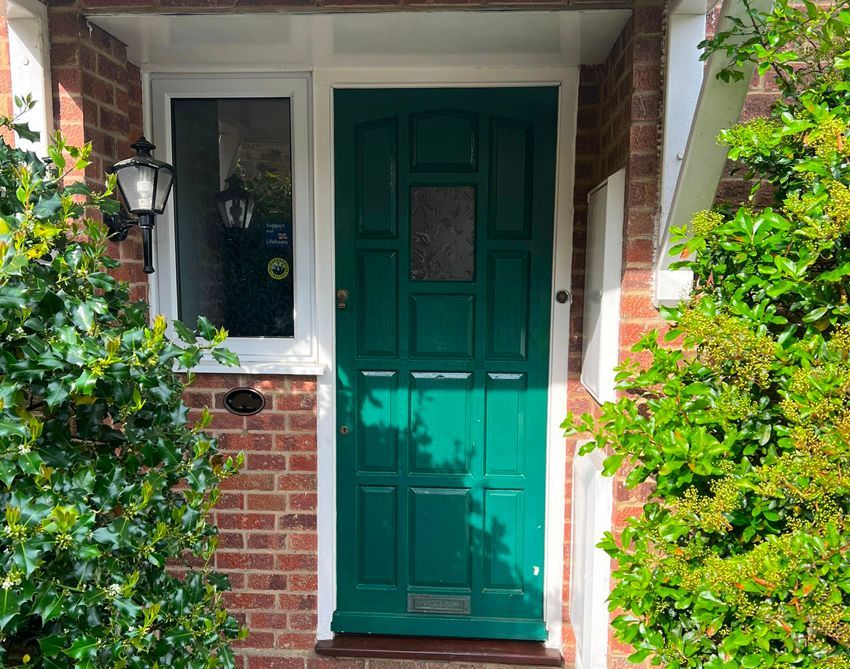 Green door with glass panel, brick surround, and side window.  Ivy and shrubbery frame the entrance.