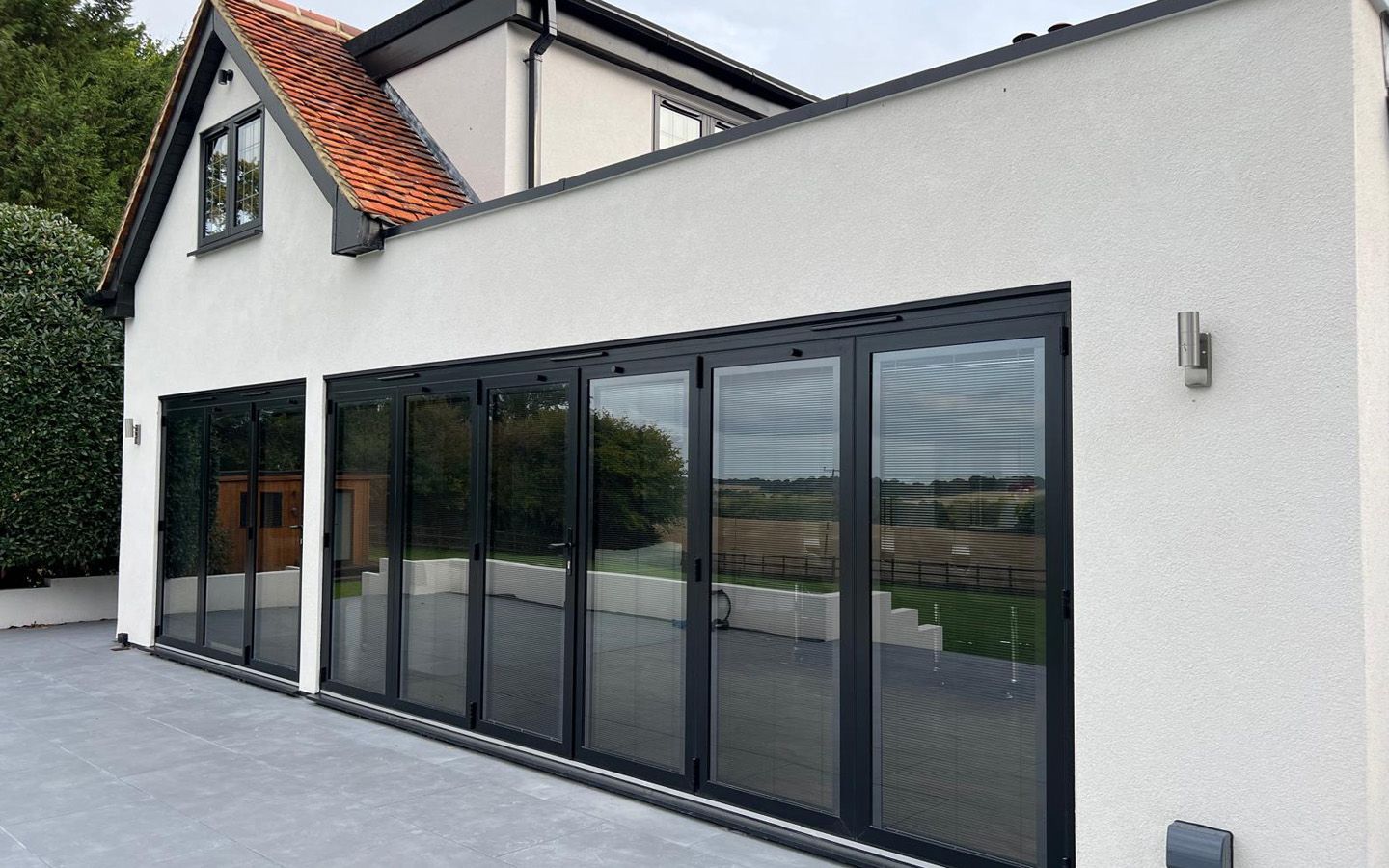Modern house with gray bi-fold doors, white walls, and red tile roof, overlooking a green field.