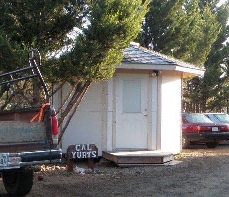Small portable white restroom beside a pickup truck in a wooded outdoor area, with a red car in the background
