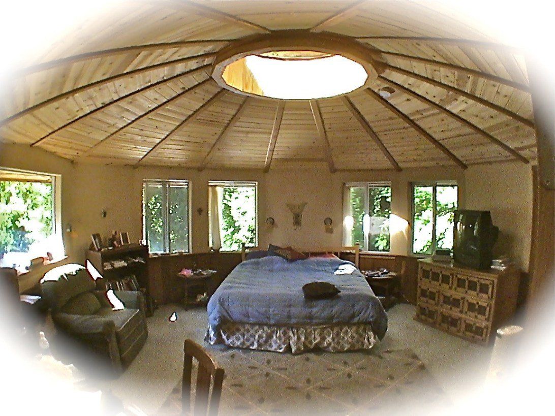 Yurt-style room with a central bed, skylight ceiling, windows, and rustic furniture