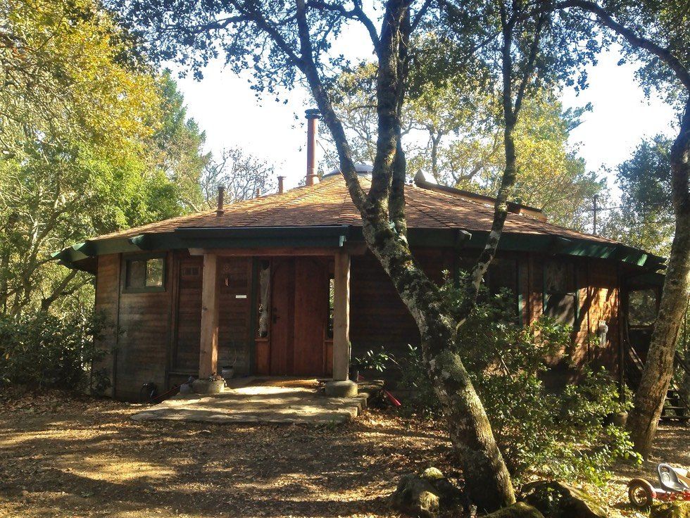 Small wooden cabin in a wooded area, with trees in front and sunlight filtering through the leaves.