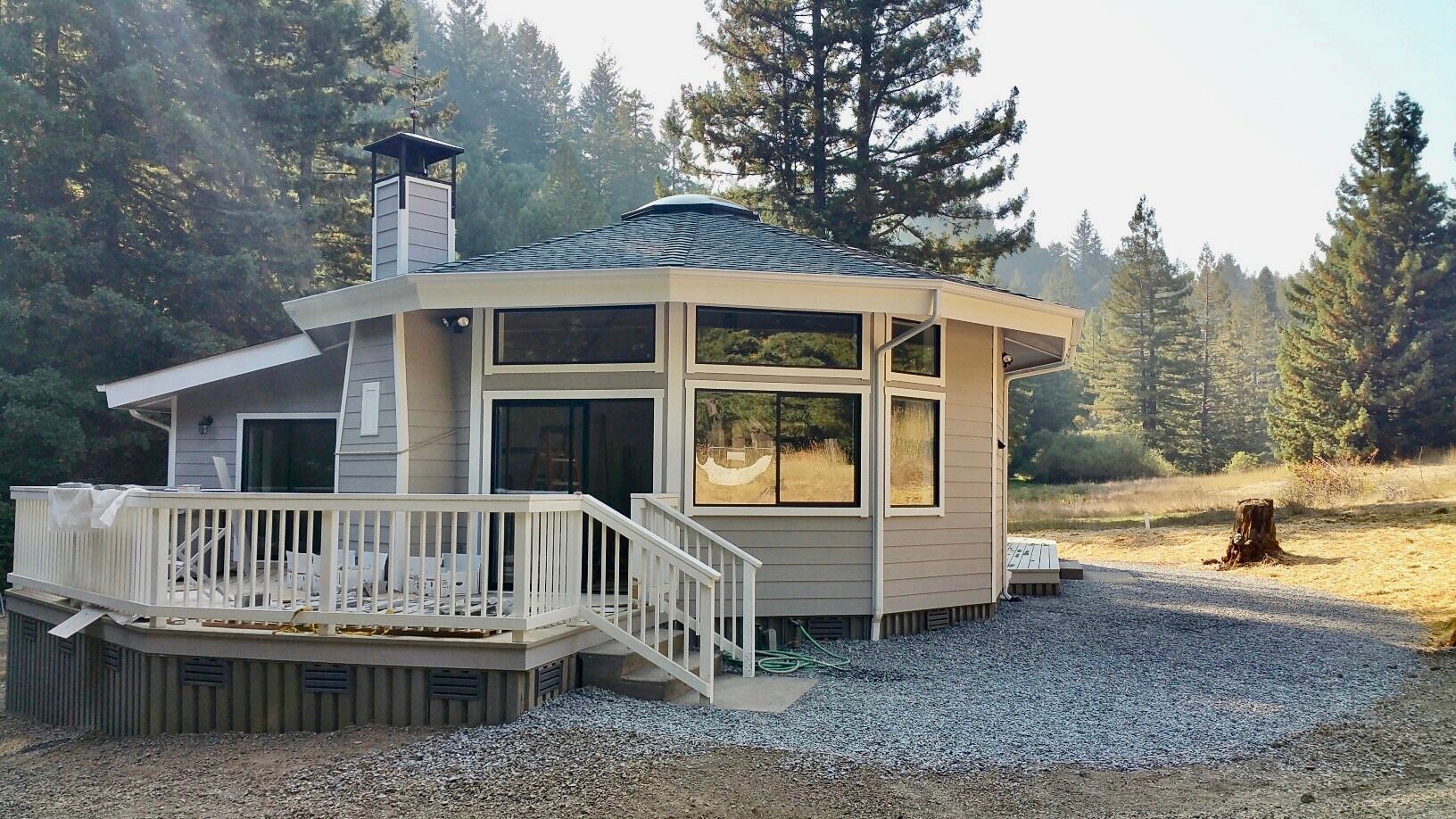 Small beige cabin with white porch in a gravel clearing, surrounded by pine trees at sunrise.