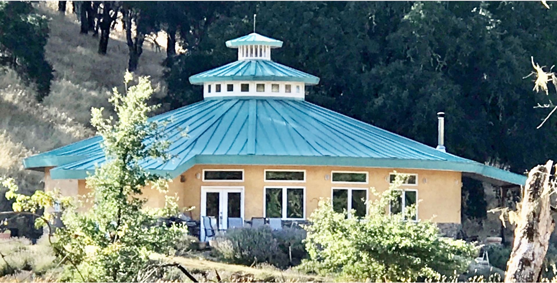 Small yellow building with teal roof and cupola, surrounded by trees and shrubs.