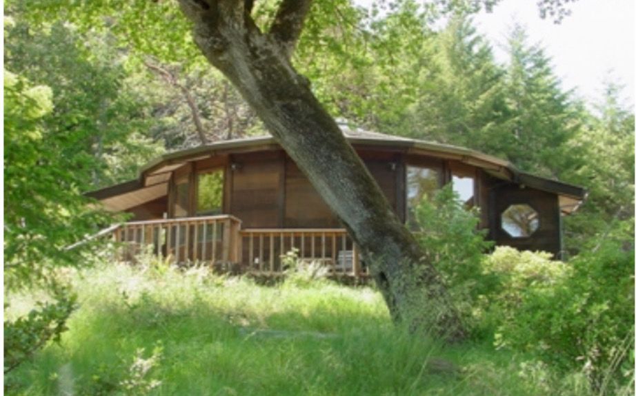 Wooden cabin in a grassy forest clearing, with a wraparound porch and slanted roof.