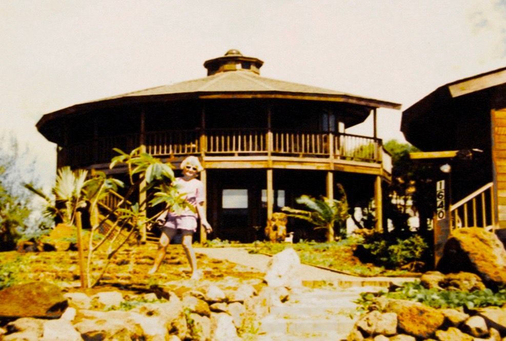 Round thatched-roof house with a wraparound balcony amid rocks and tropical plants under bright sunlight