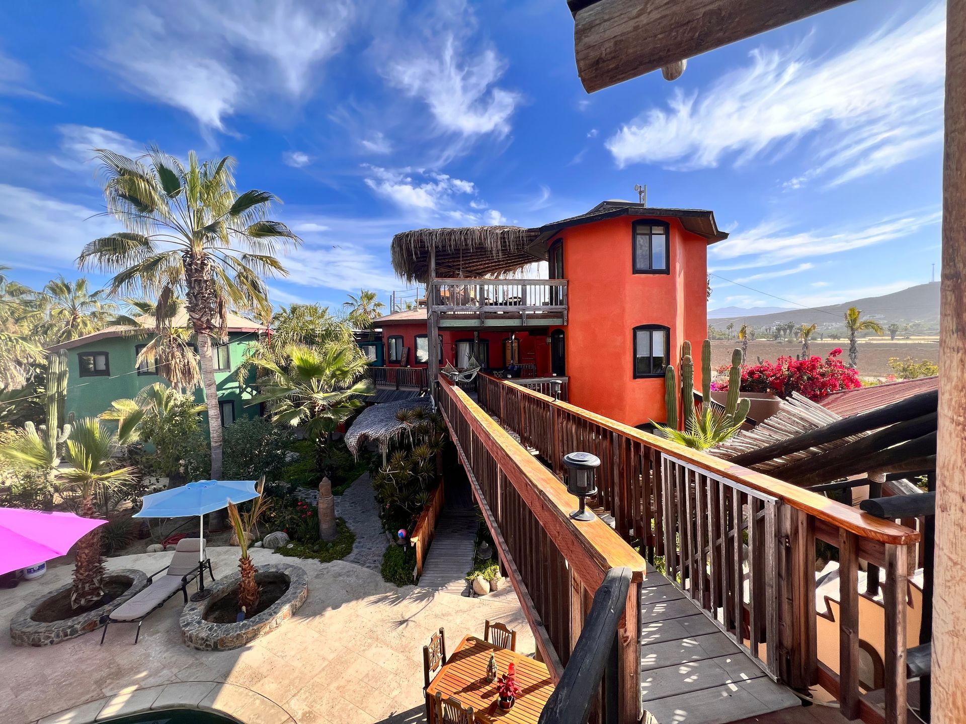 Colorful hillside resort with wooden walkway, palm trees, blue sky, and mountain view