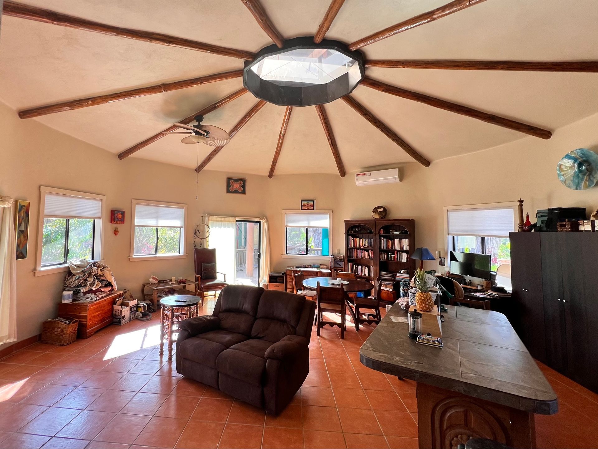 Open living room with high vaulted ceiling, brown sofa, tiled floor, windows, and kitchen island.