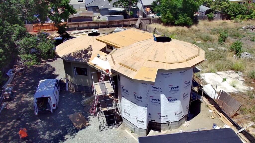 Aerial view of a round house under construction with a tan roof and white wrap, surrounded by trees and debris.