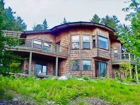 Large rustic wooden house with wraparound deck on a grassy hillside, surrounded by trees.