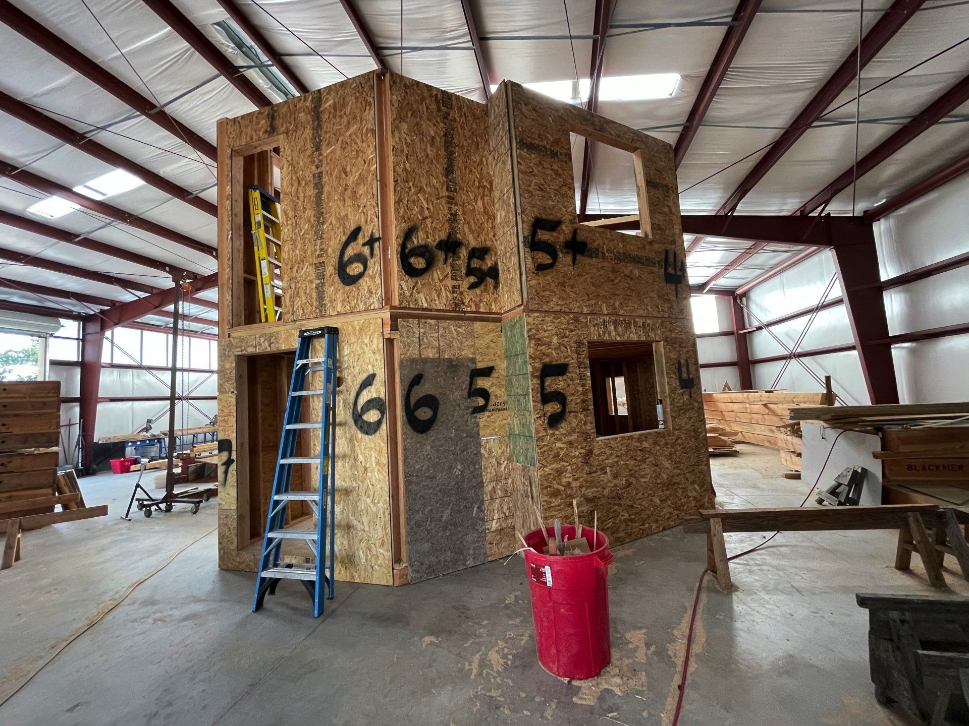 Framed house under construction with OSB walls, blue ladder, and red trash bin inside a metal shop.