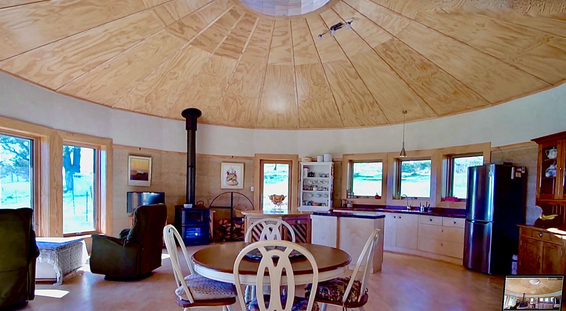 Wide-angle view of a cozy kitchen and dining area with a round wooden ceiling, windows, and a table set in the foreground