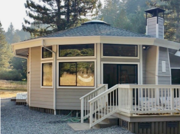 Small octagonal house with beige siding, large windows, and a white deck railing in a wooded setting