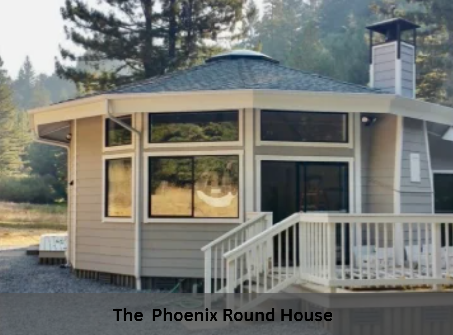 Small beige round house with white porch railing and deck in a wooded setting, labeled