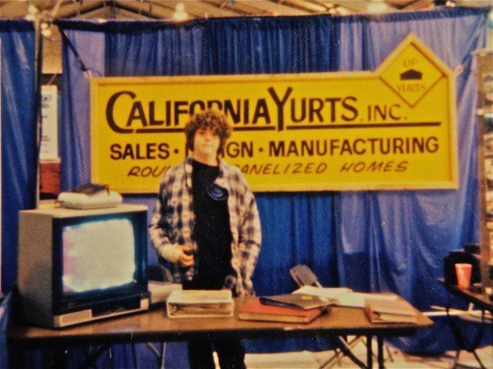 Person standing at a booth for California Yurts, Inc., with a yellow sign and blue curtains behind them