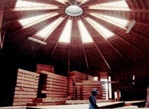 Interior of a circular wooden hall with a skylight, stacked lumber, and a person standing near the center.