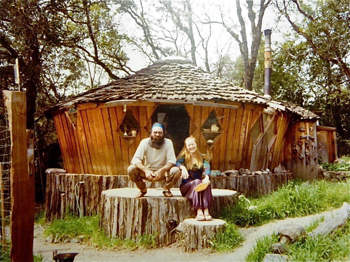 Two people sitting outside a rustic wooden hut in a wooded area