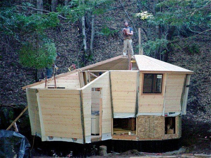 Wooden cabin under construction in a wooded area, with a person standing on the roof.