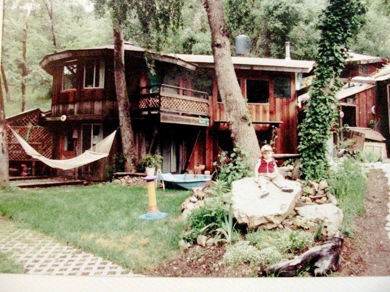 Red two-story wooden house in a wooded yard with hammock, trees, and garden seating