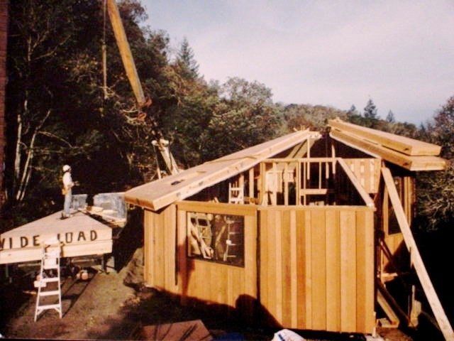 Wood-framed house under construction beside a roadside sign, with ladders and workers in a wooded area.