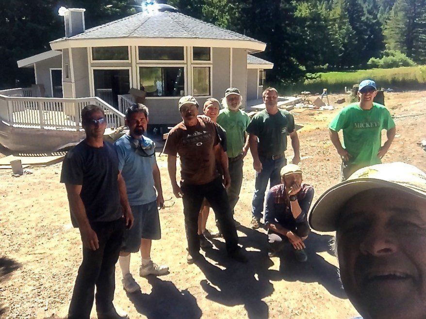 Group of people posing outdoors in front of a small building, with one person taking a selfie in the foreground.