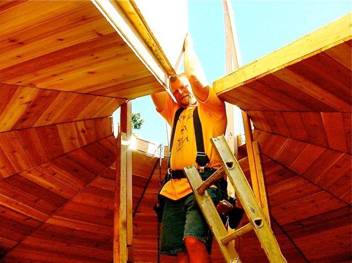 Worker on a ladder inside a wooden structure, viewed from below, with bright orange and yellow beams.