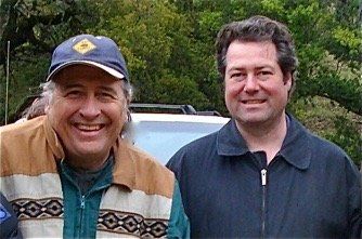 Two smiling men standing outdoors, one in a blue cap and tan jacket, the other in a dark jacket.