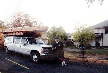 White utility truck with ladder rack parked by a person and small dog in a suburban yard after rain.
