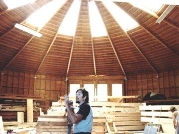 Person holding a camera inside a wooden round building with a high skylit ceiling