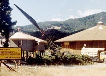 Large crane lifts a long beam beside a rural house and truck in a mountain setting.