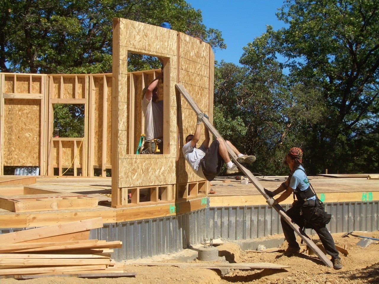 Workers framing a house wall on a construction site, lifting lumber beside a partially built wooden structure.