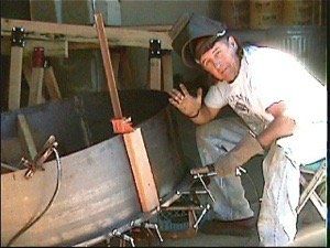 Person in a workshop kneels beside a large metal object, smiling and waving.